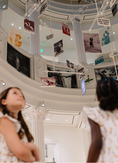 Two girls look up at a museum display of hanging photographs and art inside a large, white, column-filled rotunda. The images are suspended above, and the space feels bright and modern.