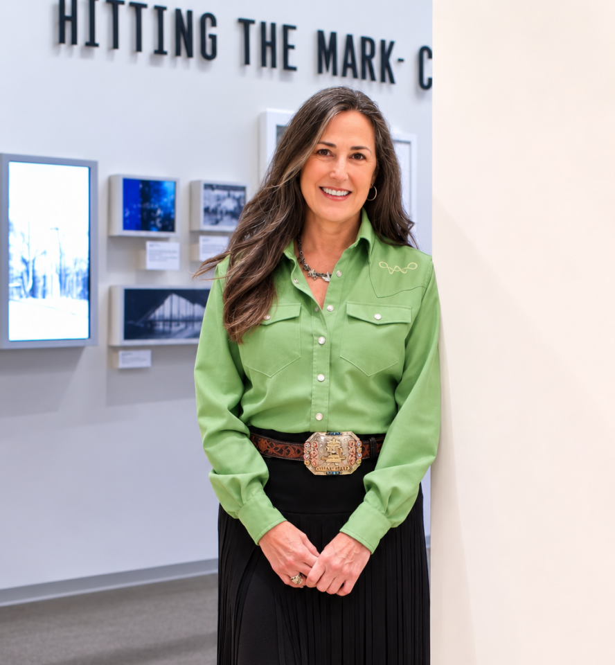 Smiling woman in a green western-style shirt and decorative belt buckle, standing in a gallery with framed photos.
