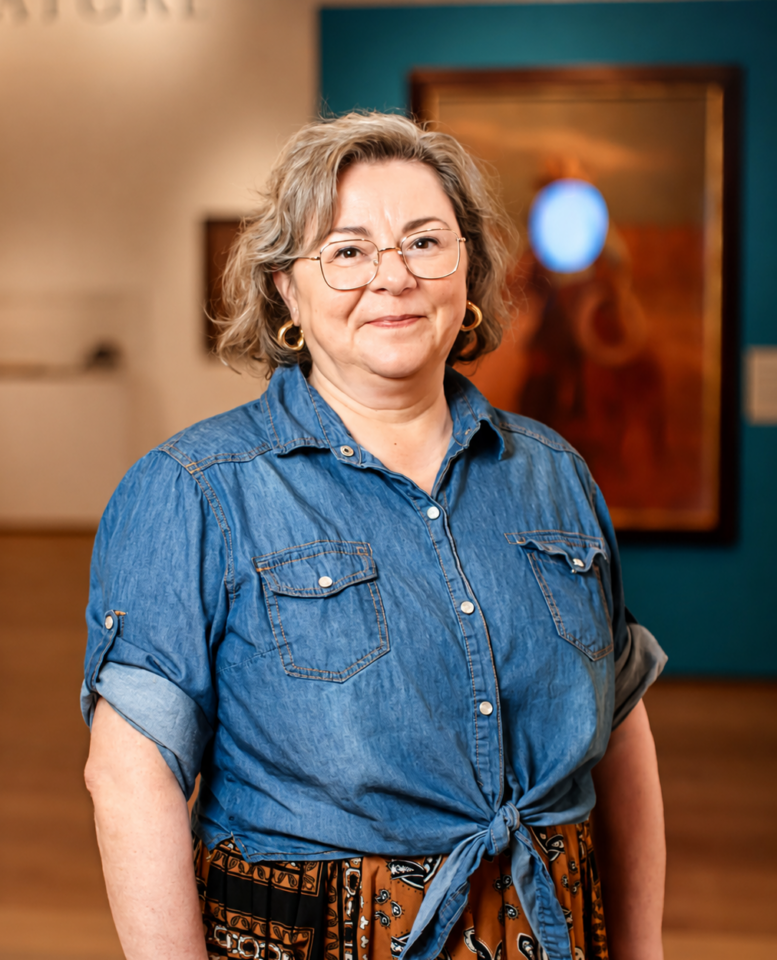 Woman with short gray hair and glasses stands in an art gallery wearing a denim shirt and patterned skirt.