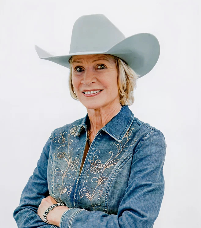 Woman wearing a light gray cowboy hat and embroidered denim shirt, smiling with arms crossed against a plain white background.