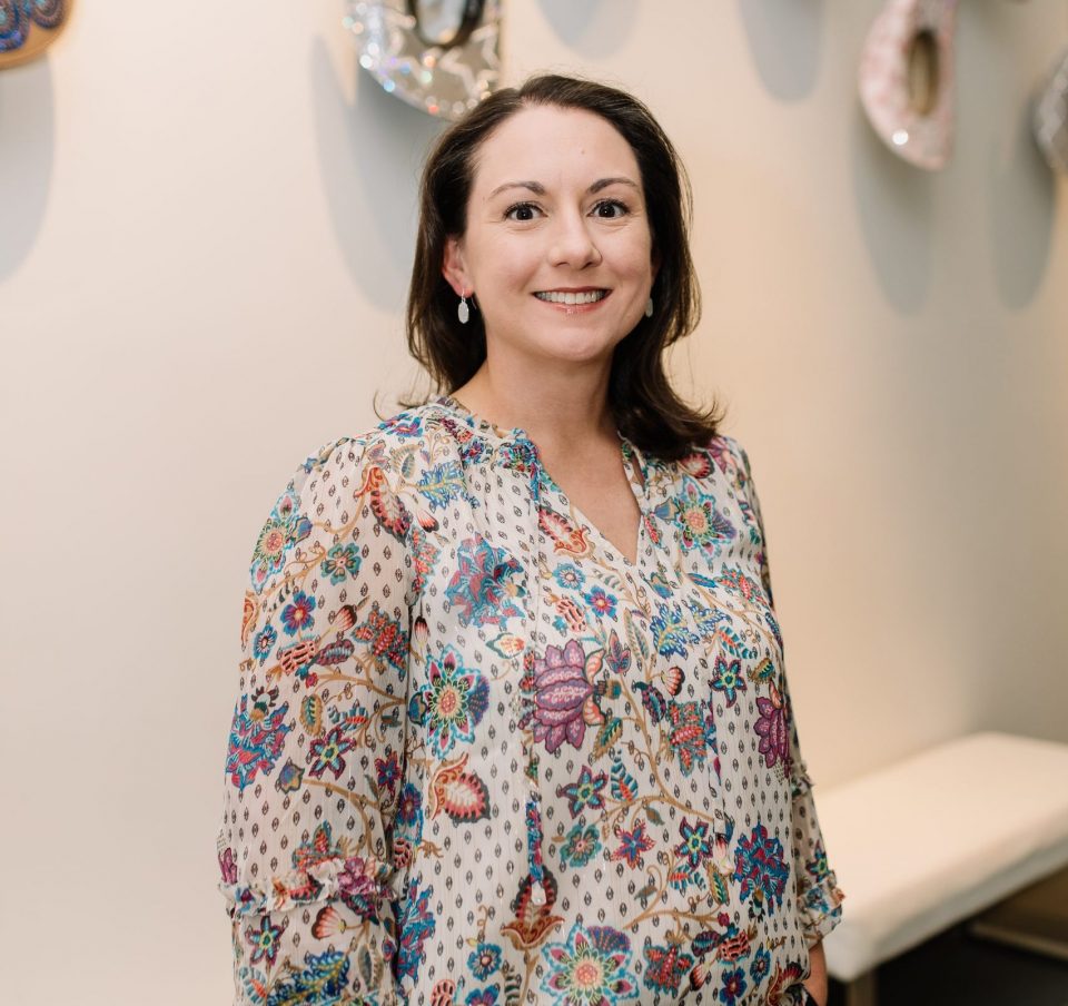 Smiling woman in a white, colorful floral blouse standing indoors with her hands in her pockets.