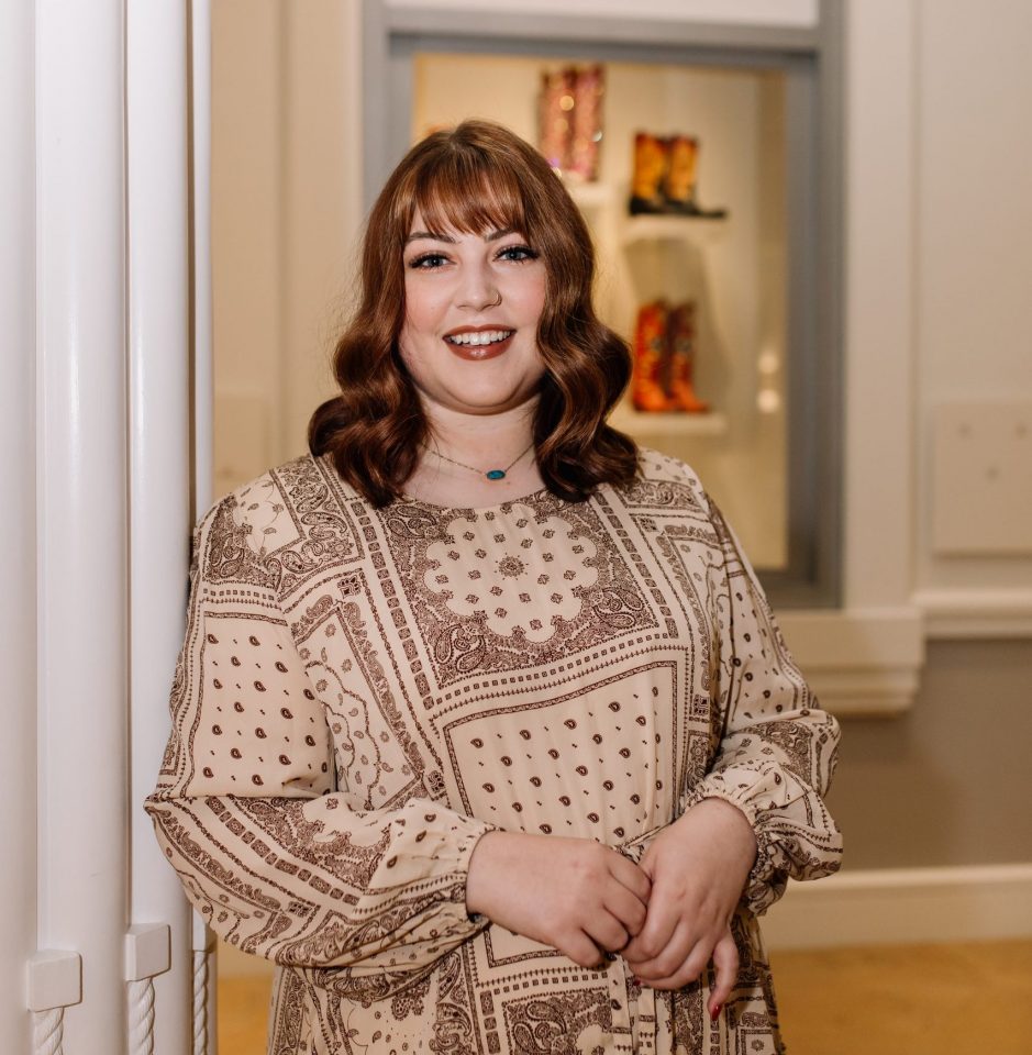 Smiling woman with brown wavy hair and a nose ring wearing a beige paisley dress, standing by a doorway indoors.