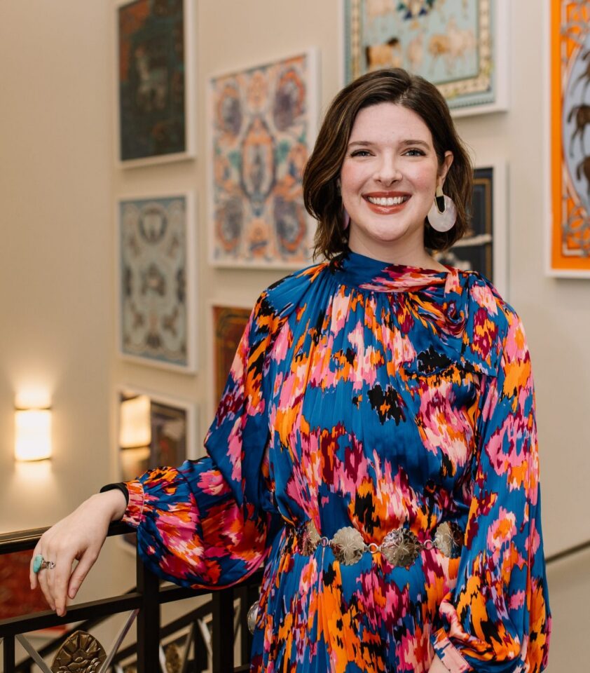 Smiling woman in a bold, multicolor abstract-print dress stands near a black railing in an art gallery with framed artworks in the background, pink earrings visible.