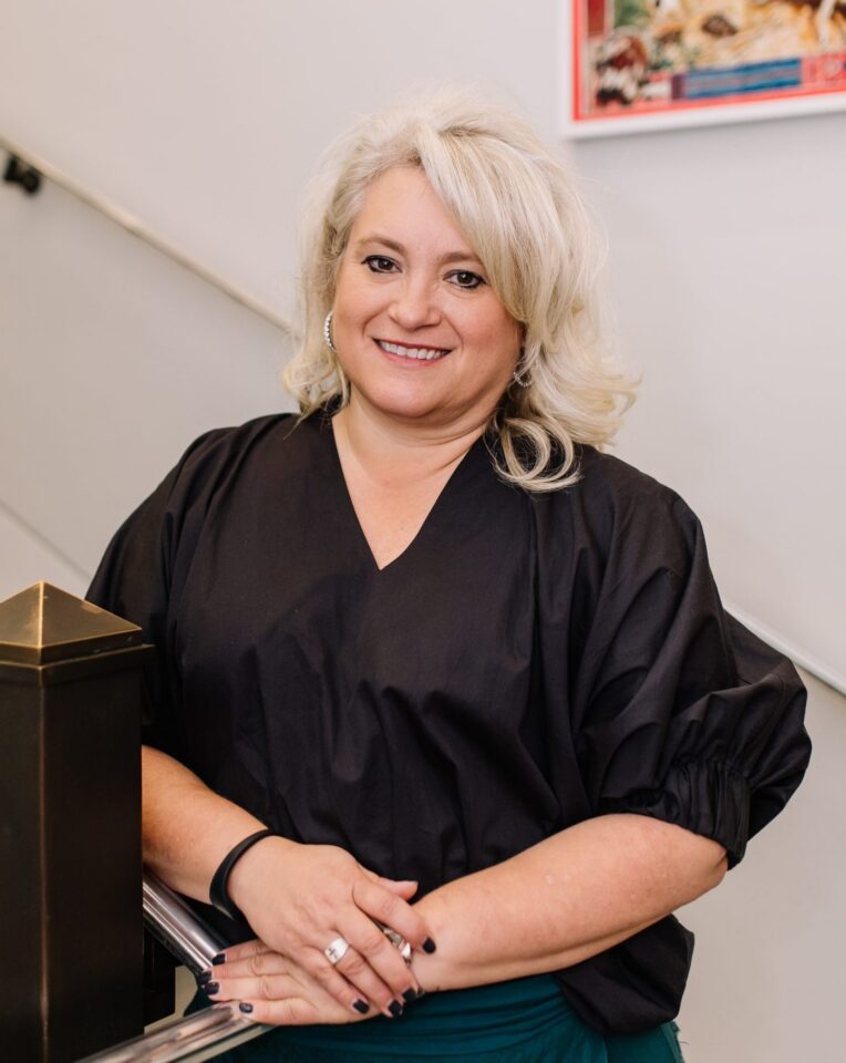 Smiling woman with short blonde hair in a black blouse leaning on a railing beside a staircase in a light-filled setting.