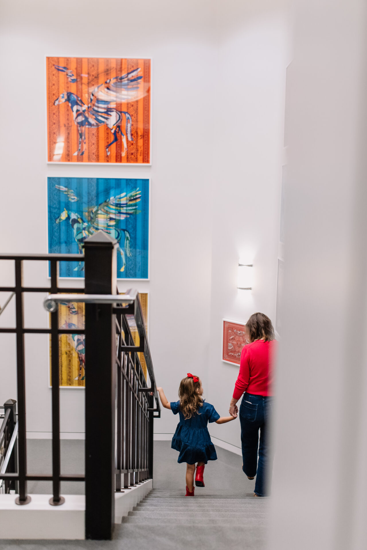 A child in a blue dress and red boots holds hands with an adult while walking up stairs, with colorful animal artwork hanging on the wall beside them.