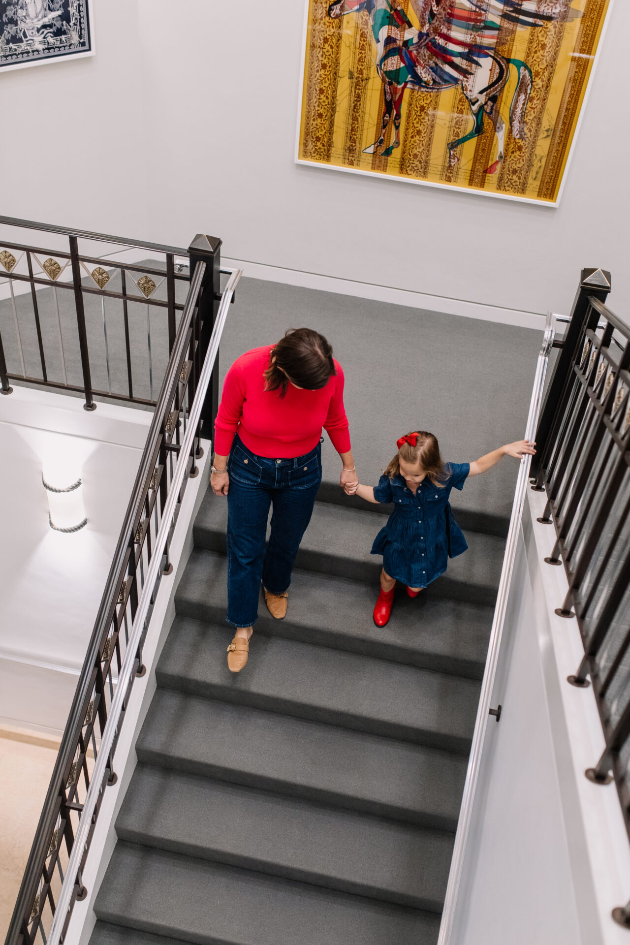 A woman in a red top holds hands with a young girl in a blue dress and red boots as they walk down a gray-carpeted staircase in a brightly lit building.