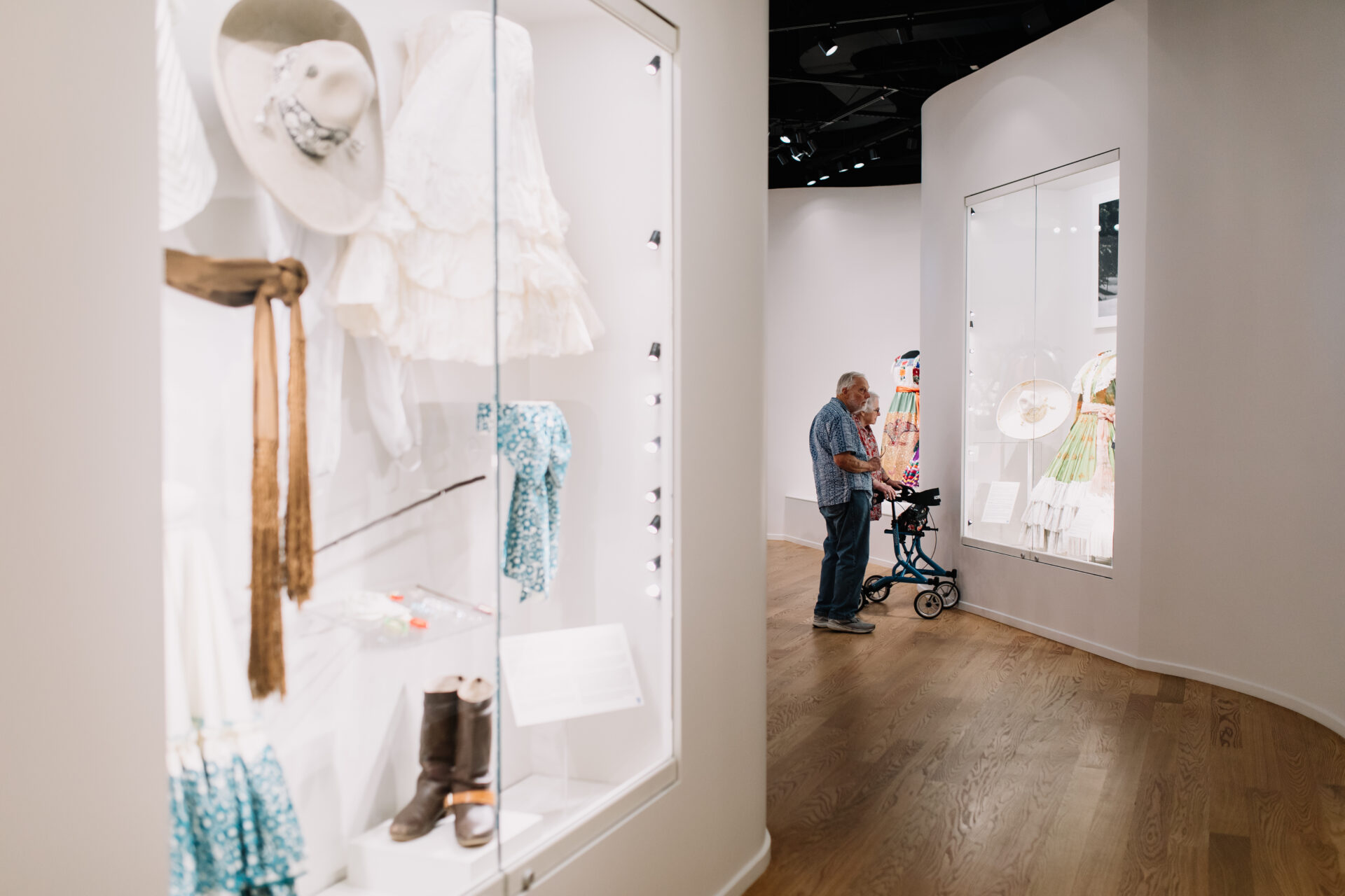 A man uses a walker while looking at fashion exhibits displayed in glass cases along a curved, white-walled hallway in a museum.