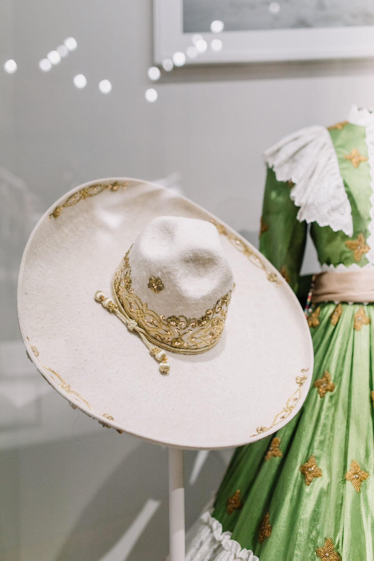 A white sombrero with gold embroidery is displayed on a stand in front of a green traditional dress with gold patterns and white lace details.