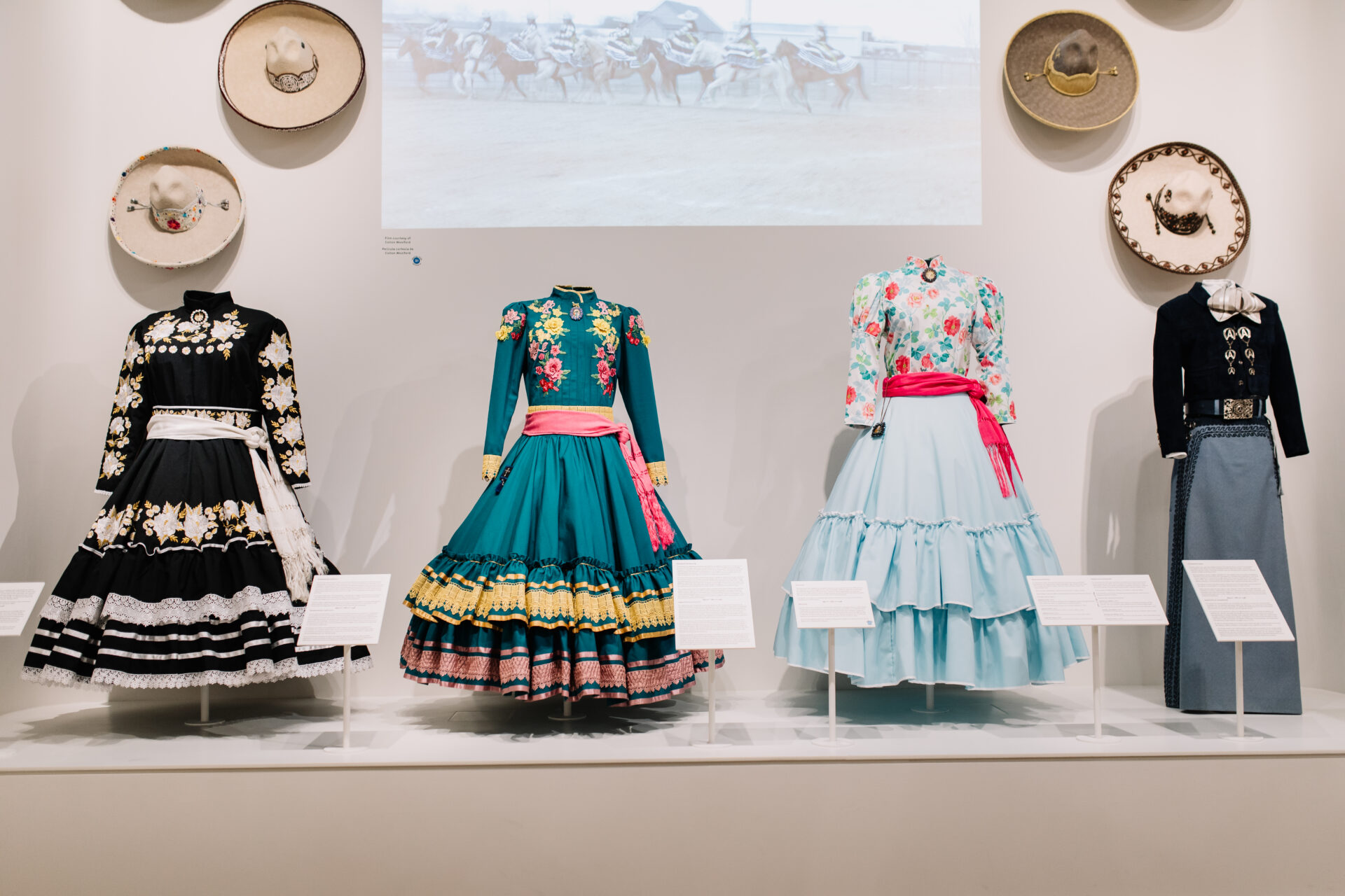 Four traditional Mexican dresses with floral embroidery and hats are displayed on mannequins in a museum exhibit, with informational placards and a projected image of horse riders in the background.