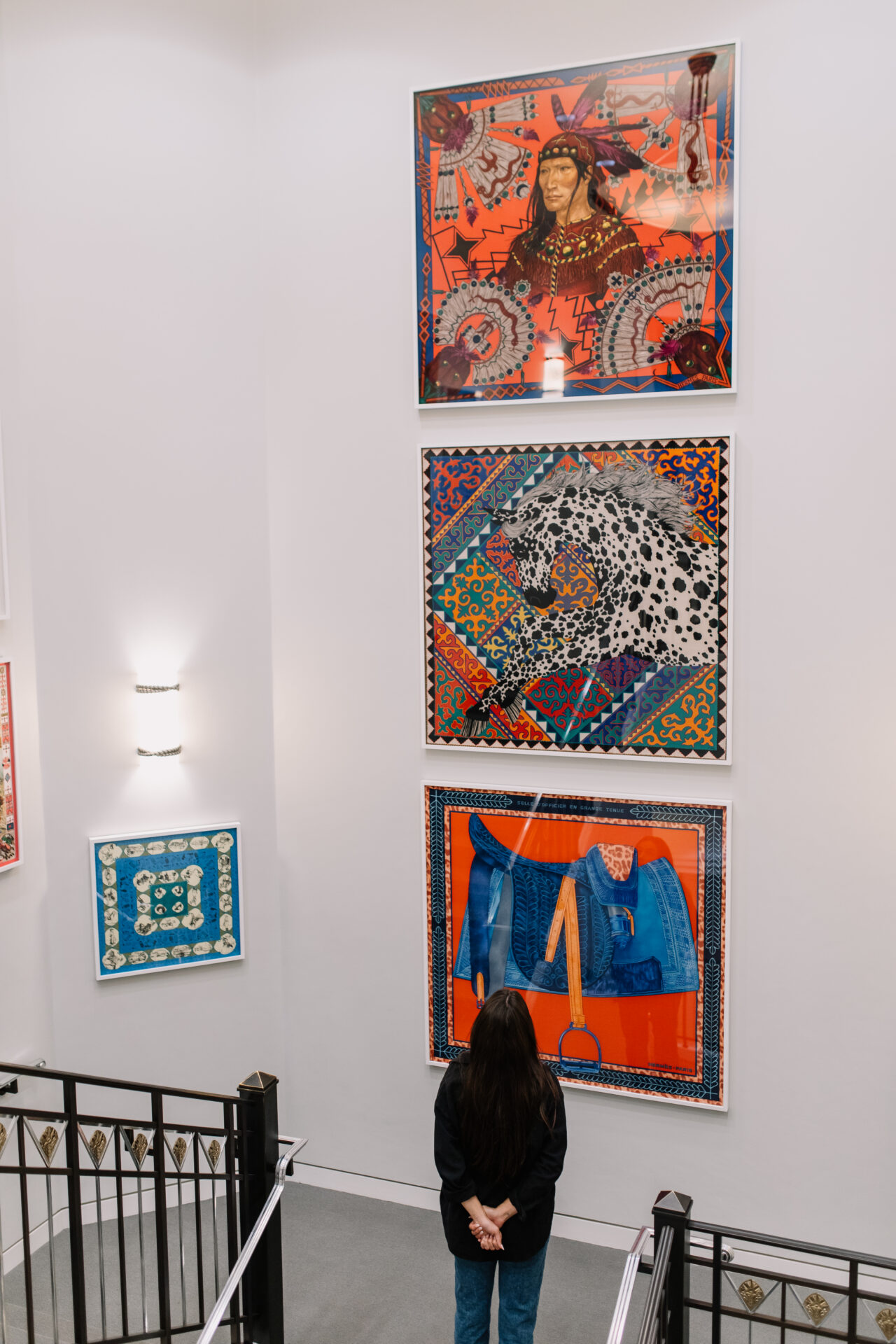 A person with long hair stands, hands behind back, looking at three colorful framed artworks on a white gallery wall, including a portrait, a leopard, and blue jeans with a handbag.