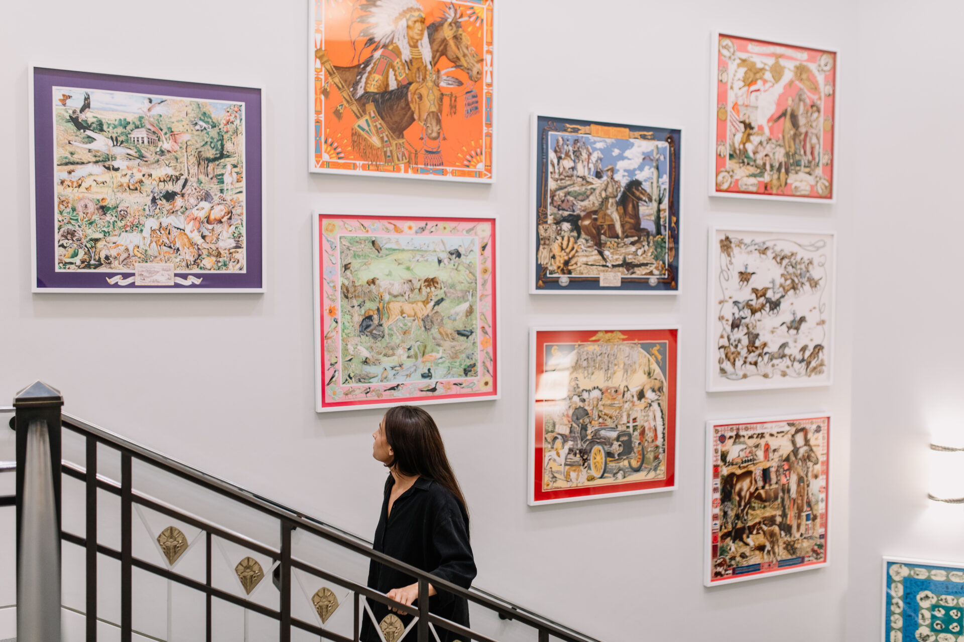 A woman walks up a staircase next to a wall displaying nine colorful, framed art pieces featuring various scenes with animals and people.
