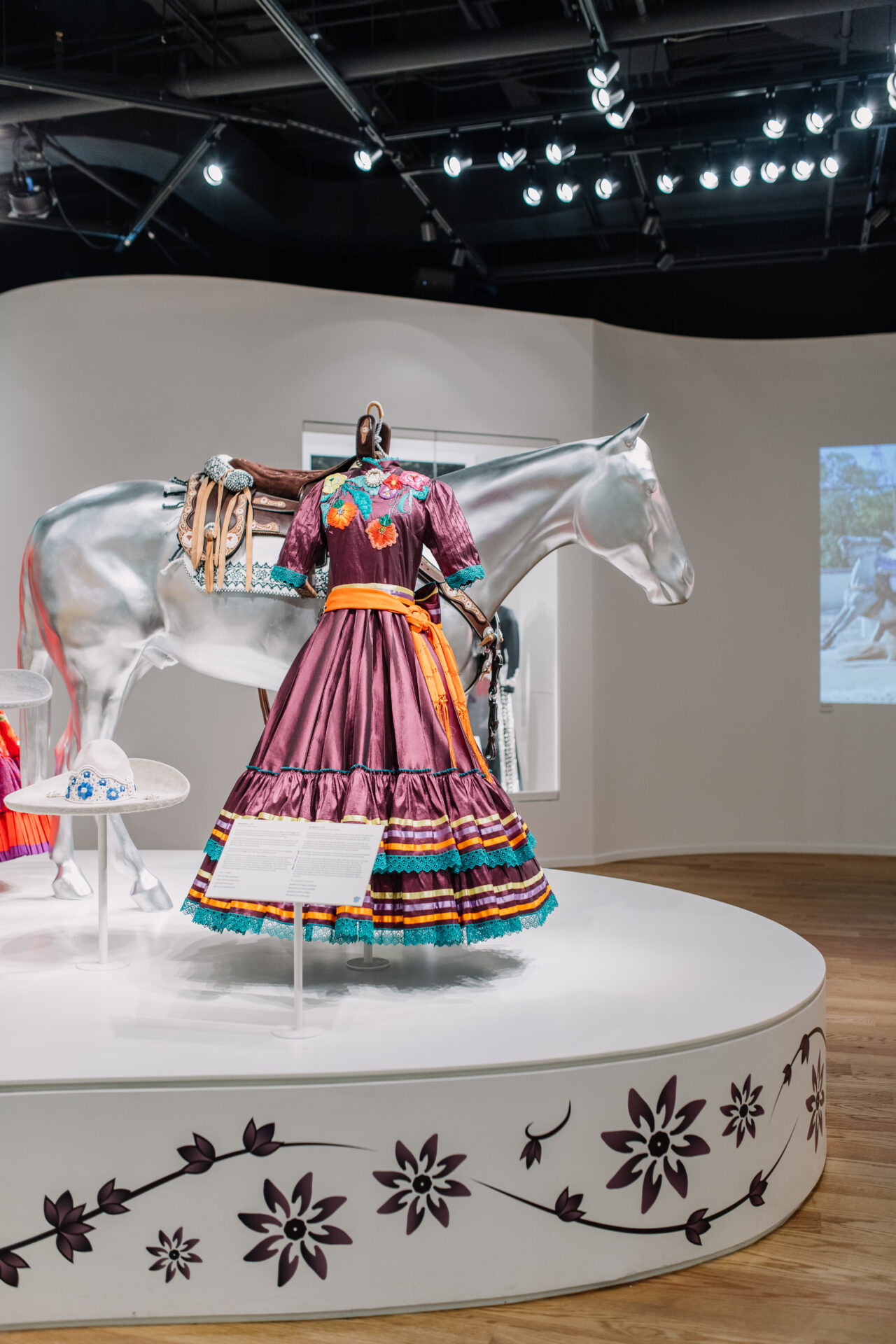 A colorful traditional dress is displayed on a mannequin next to a white horse statue with a saddle, all on a round platform with floral designs in a museum exhibit.