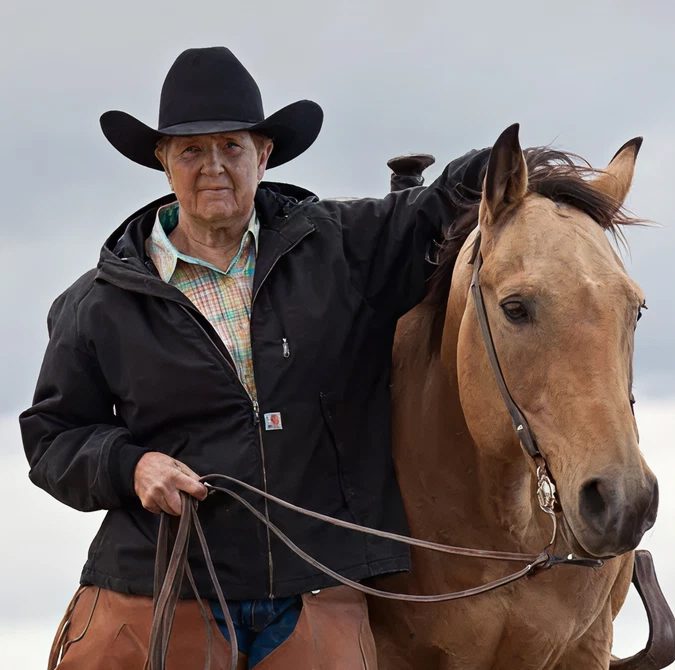 An older man in a black cowboy hat and jacket stands next to a tan horse, holding its reins. The sky is cloudy and gray.