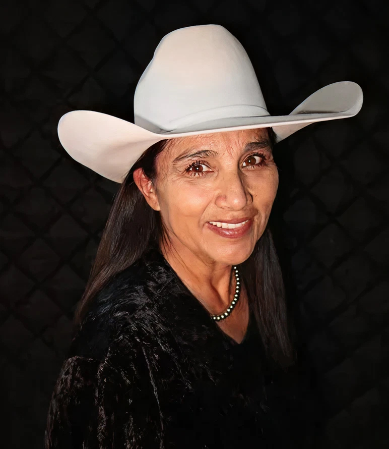 A woman with long dark hair, wearing a white cowboy hat, a black textured jacket, and a beaded necklace, smiles in front of a dark, patterned background.