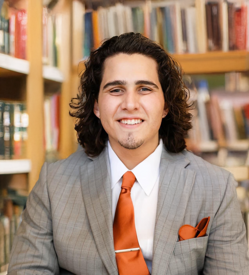 A young man with long curly hair in a gray suit and orange tie sits and smiles in a library with bookshelves in the background.