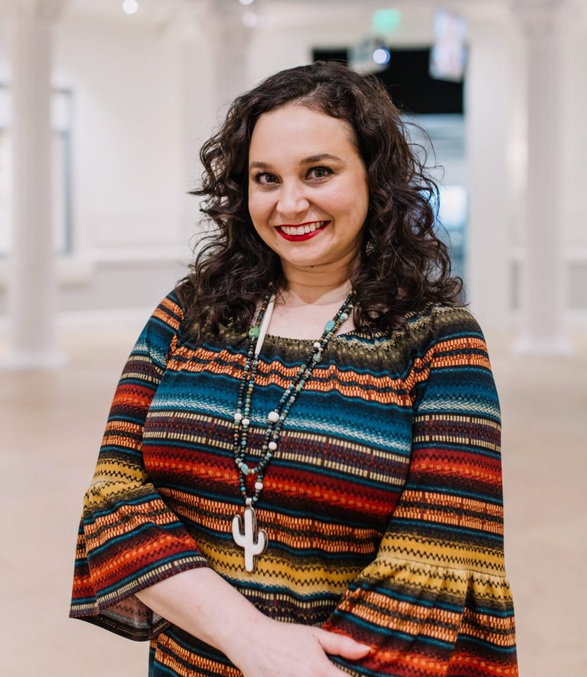 Smiling woman with wavy dark hair in a multicolored striped top and beaded necklace.
