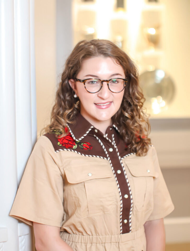 Smiling woman with curly hair and glasses standing indoors, wearing a beige shirt with brown trim and red floral embroidery.