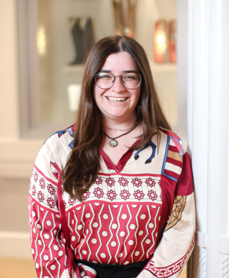 Smiling woman with long brown hair and round glasses, wearing a red and white patterned blouse indoors near a white column.
