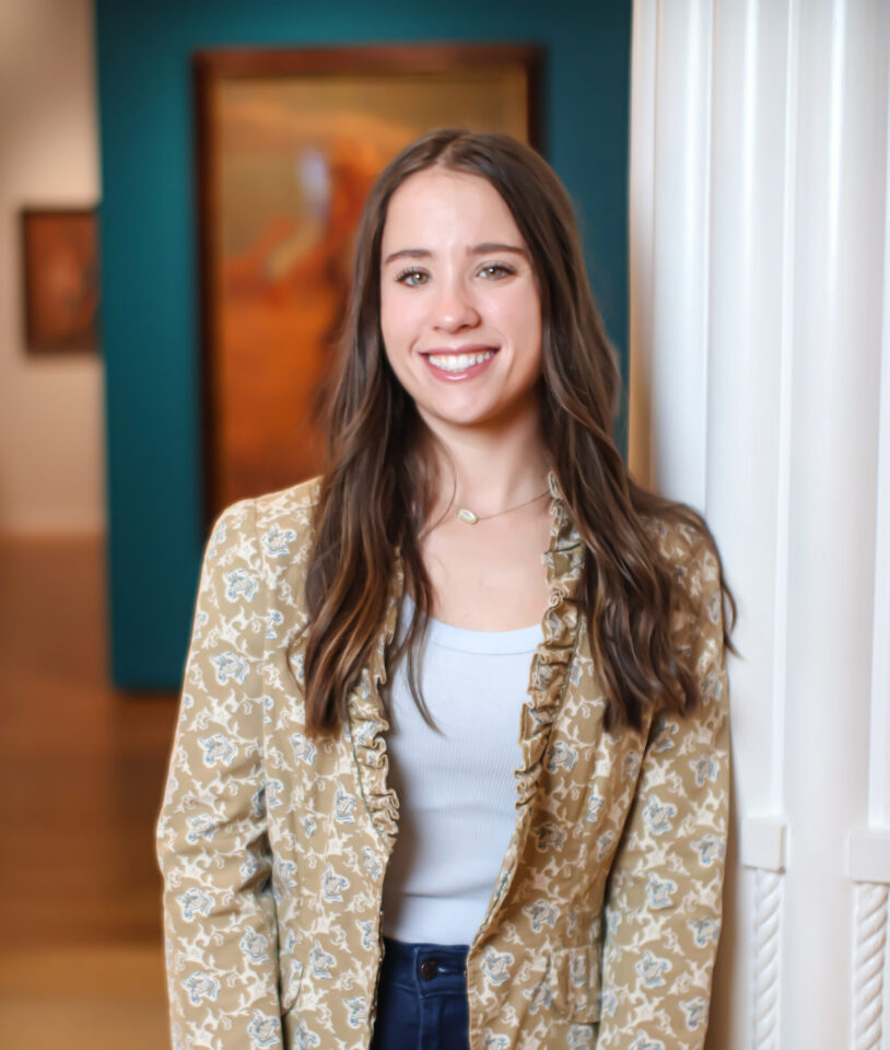 Smiling woman with long brown hair in a beige floral blazer and white top standing indoors, blurred art in the background.
