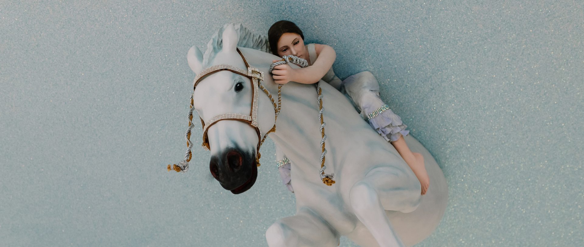 A porcelain figurine of a girl in a lavender dress hugging the neck of a white horse with gold-trimmed tack, set against a light blue glittery background.