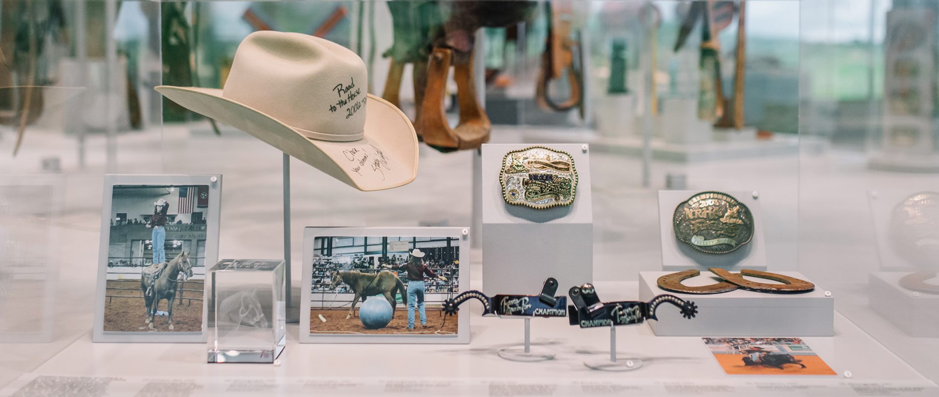 Display case with a signed cowboy hat, photos of rodeo events, a belt buckle, riding spurs, horseshoes, and a large commemorative coin, all related to western or rodeo culture.