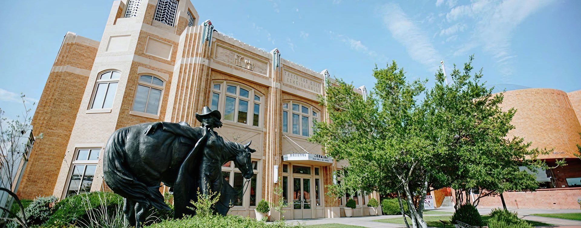 A large bronze statue of a cowboy herding longhorn cattle stands in front of a historic brick building with arched windows and trees, under a blue sky.