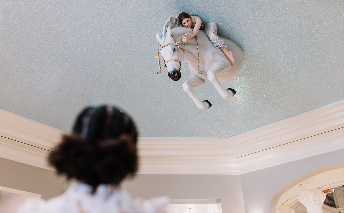 A person in the foreground looks up at a ceiling where a sculpture of a girl riding a white horse is mounted upside down. The ceiling and sculpture are brightly lit.