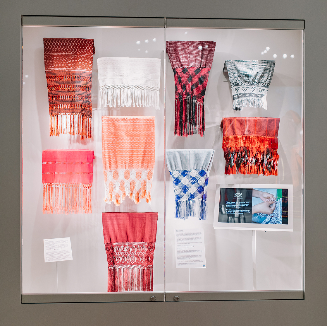 Nine intricately woven sashes with various patterns and fringe hang in a glass display case. An informational plaque and a small screen are displayed on the lower right side. The background is white.