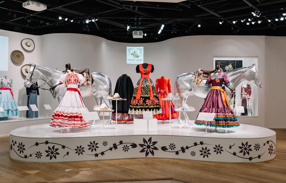 Colorful traditional dresses are displayed on mannequins between two silver horse statues in a well-lit exhibit. Embroidered hats and accessories are arranged on the platform, with floral designs decorating the base.