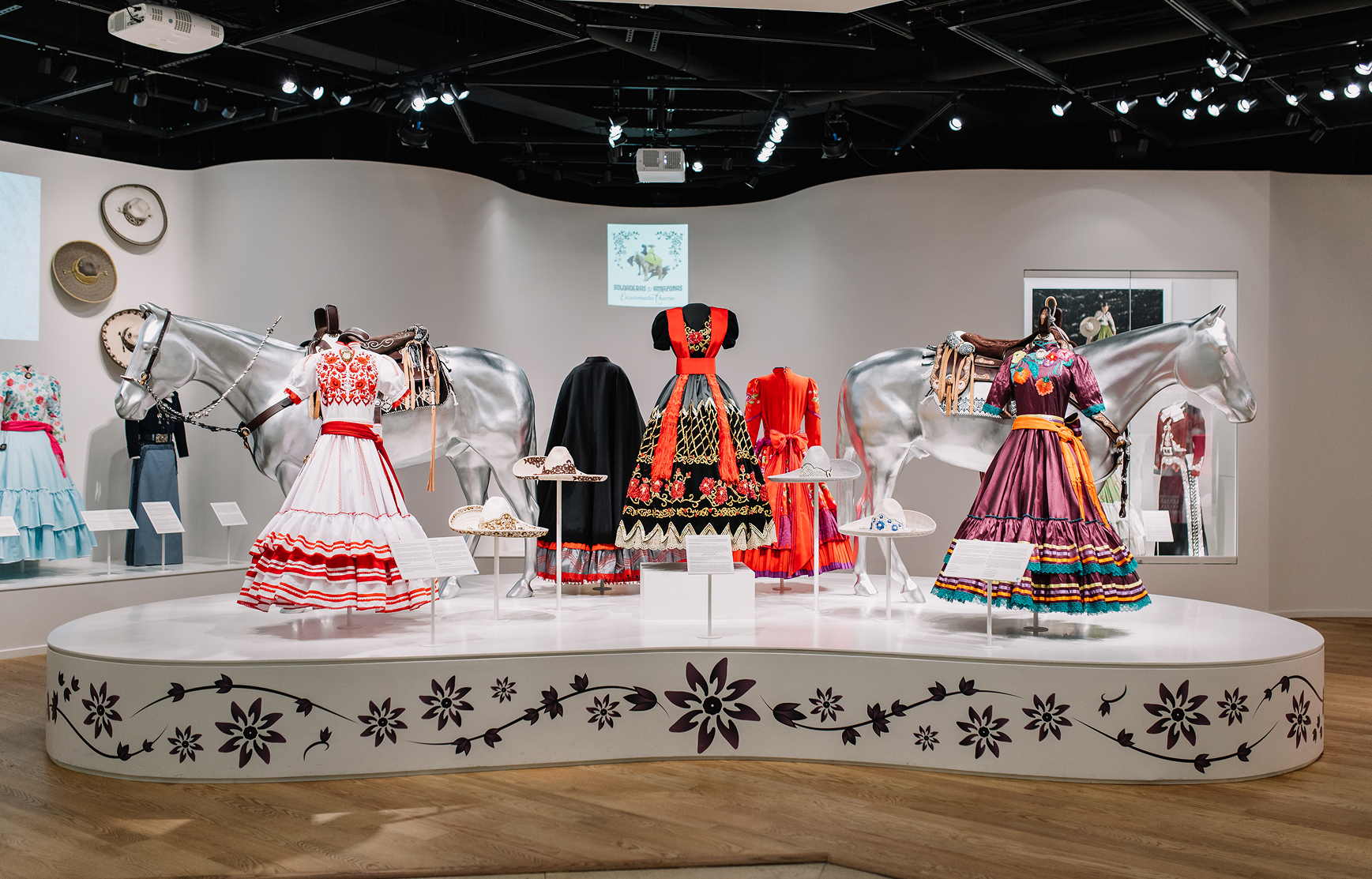 Colorful traditional dresses are displayed on mannequins between two silver horse statues in a well-lit exhibit. Embroidered hats and accessories are arranged on the platform, with floral designs decorating the base.