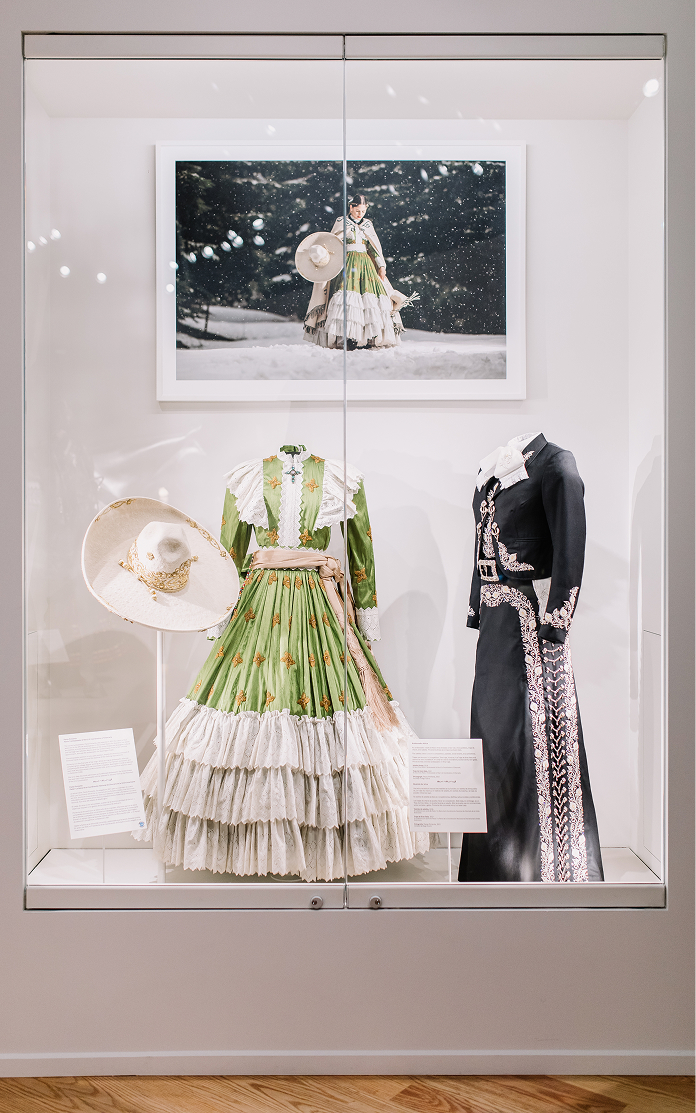 Two traditional Mexican outfits, one green and white with a wide skirt and hat, and one black with silver details, displayed in a glass case with a matching photo above them.