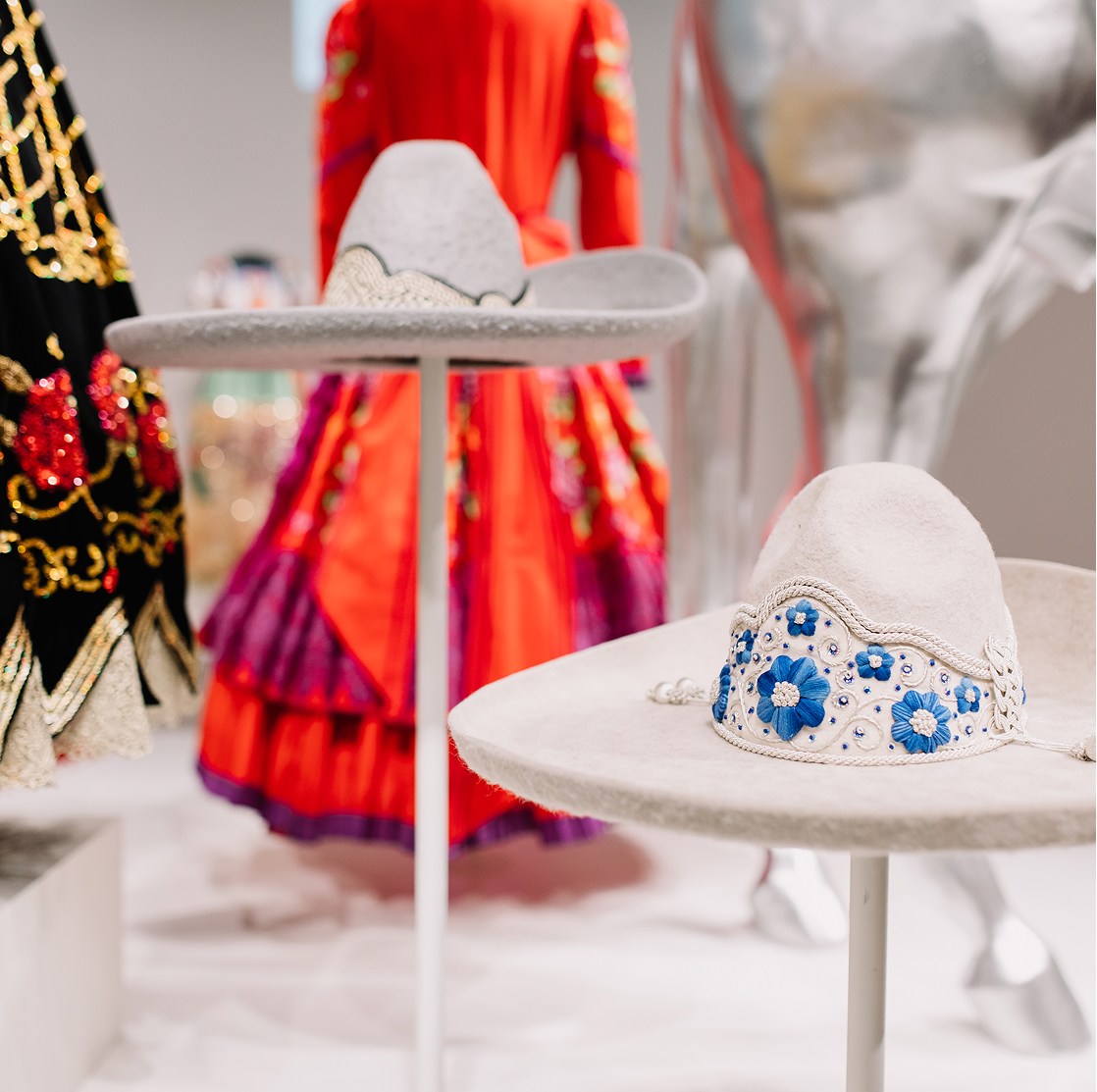 Two ornate white sombreros with blue floral embroidery are displayed on stands. In the background, theres a bright red traditional dress and a silver horse sculpture, slightly out of focus.