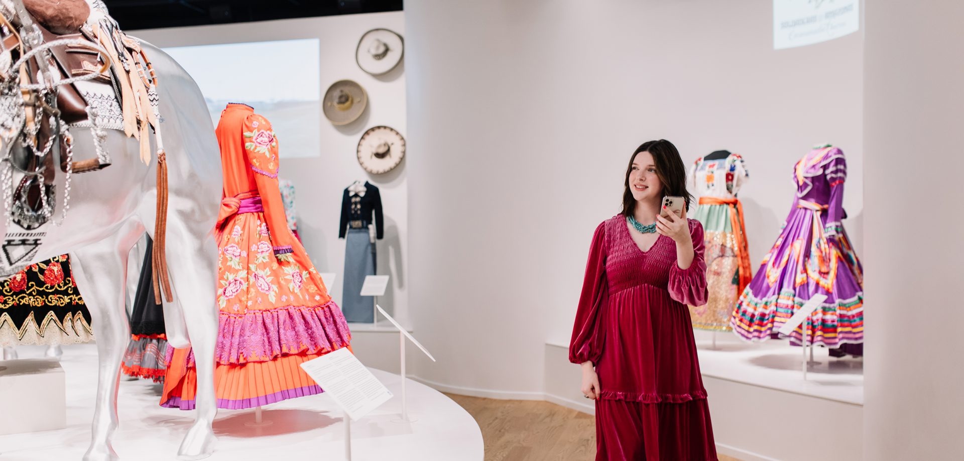 A woman in a red dress stands in a museum exhibit of colorful, traditional dresses and a white horse statue, admiring the display and holding her phone.