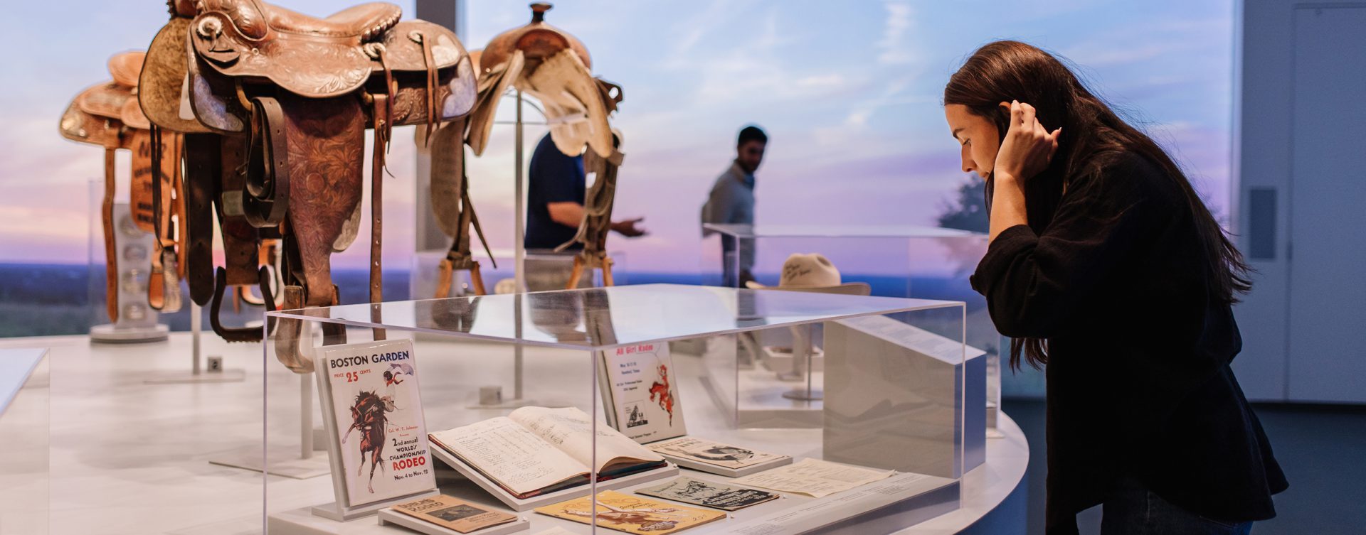 A woman looks closely at books and old posters in a glass display case at a museum, with horse saddles and two other people in the background.