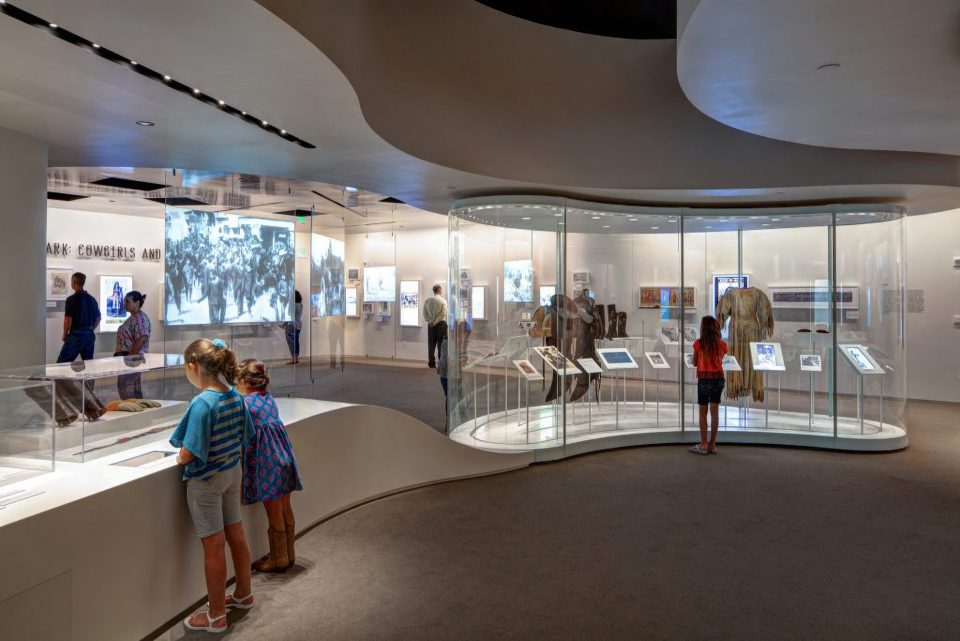 A museum gallery with curved glass displays, historical clothing, and artifacts. Several children and adults view exhibits. Digital screens and historic photos are visible on the walls.