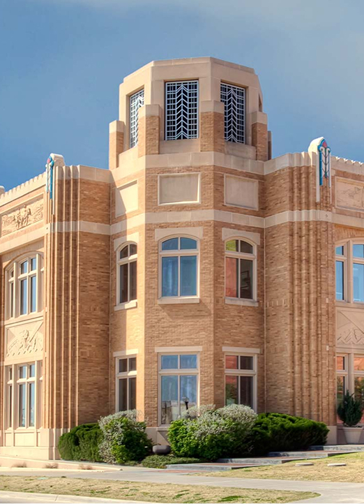 A tan brick building with Art Deco design, featuring tall vertical accents, large windows, and an octagonal tower with decorative metalwork at the top, surrounded by shrubs and a blue sky.