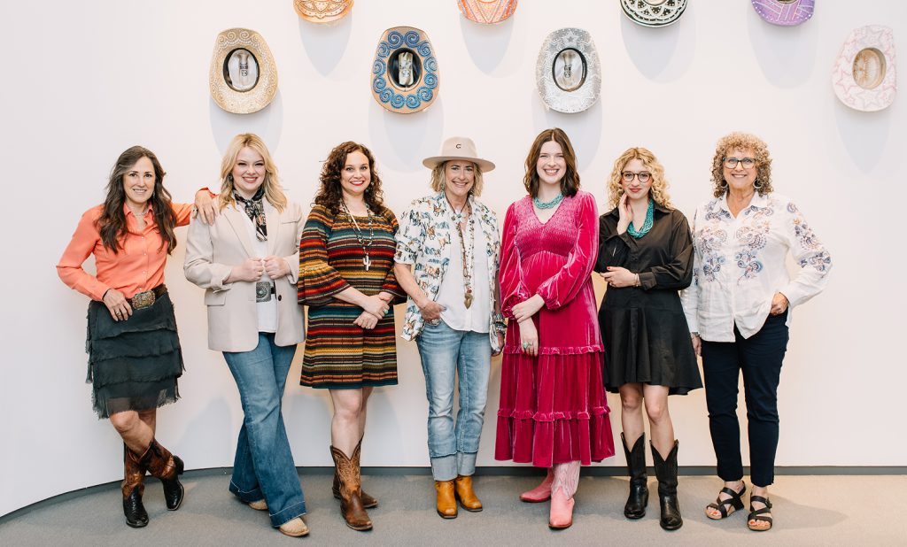 Seven women pose together in front of a white wall adorned with colorful, decorative pottery pieces. They wear varied stylish outfits, including dresses, boots, and jewelry, and all are smiling at the camera.
