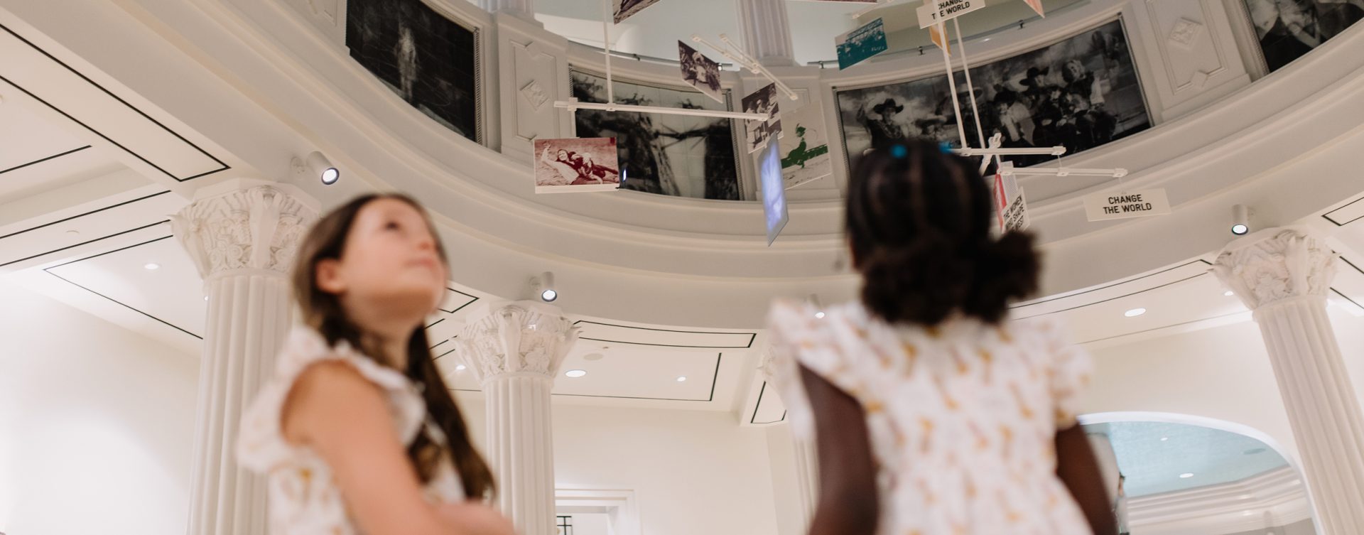 Two young girls in patterned dresses look up at hanging art and photos on display in a white, columned museum with a domed ceiling.