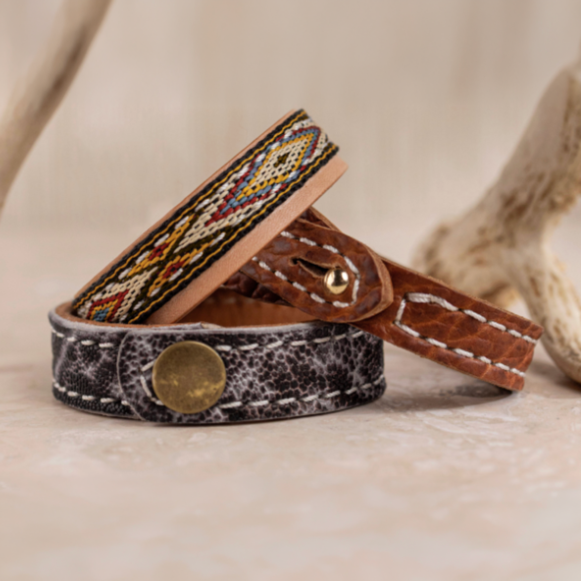 Two handmade leather bracelets, one with colorful beadwork and the other with a distressed finish and brass snap, displayed upright on a light surface near antler pieces.