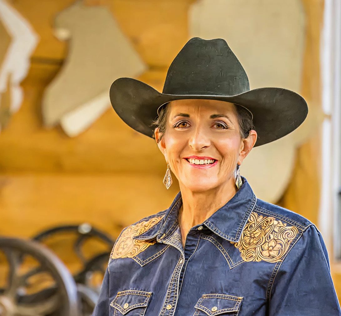 A smiling woman in a black cowboy hat and blue embroidered shirt stands indoors with western decor in the background.