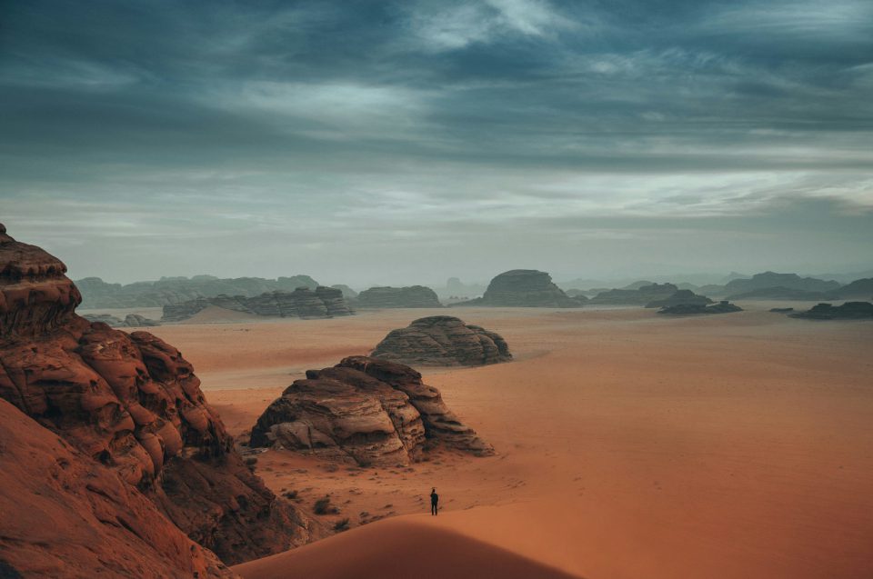 A person stands on a sand dune in a vast desert landscape with scattered rock formations under a dramatic cloudy sky.