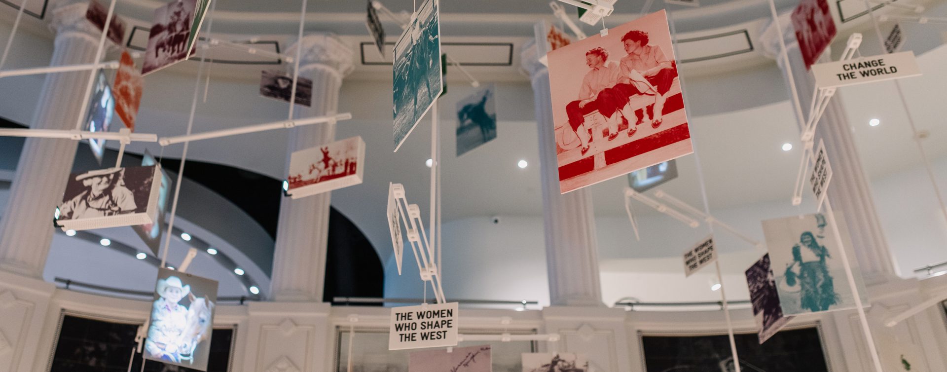 Multiple photographs and signs, including THE WOMEN WHO SHAPE THE WEST and CHANGE THE WORLD, hang from white rods in a bright, columned room.