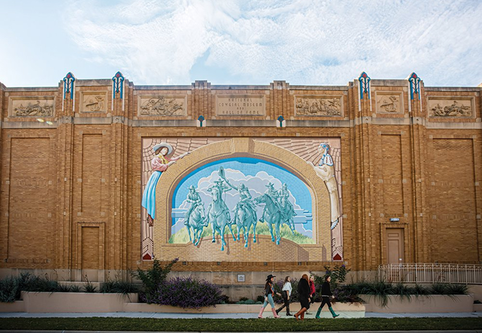 Four people walk past a large tan brick building featuring a colorful mural of cowboys riding horses under an archway, with blue sky and clouds above.