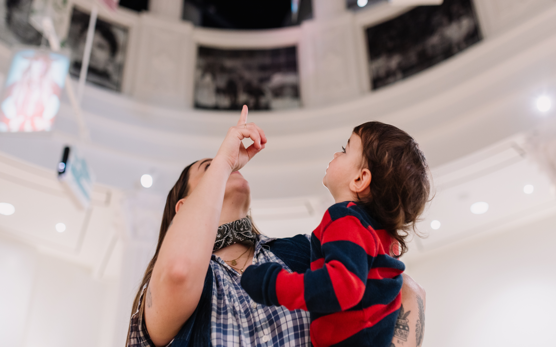 A woman holds a young child in her arms while pointing upward. The child looks up, following her gesture. They are indoors, possibly in a museum or gallery with white walls and circular architecture.