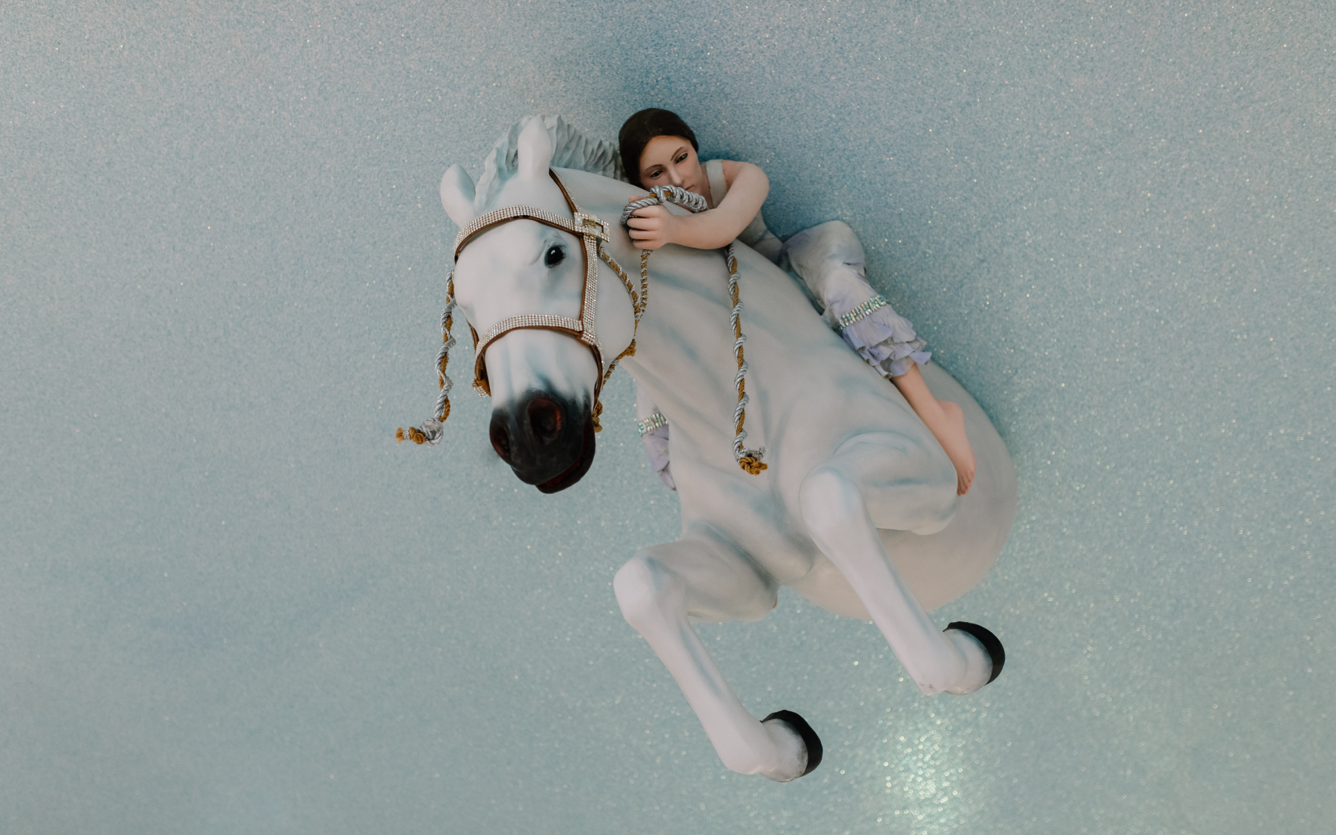 A figurine of a woman in a light purple dress clings to the neck of a rearing white horse with gold braided reins, set against a sparkly light blue background.