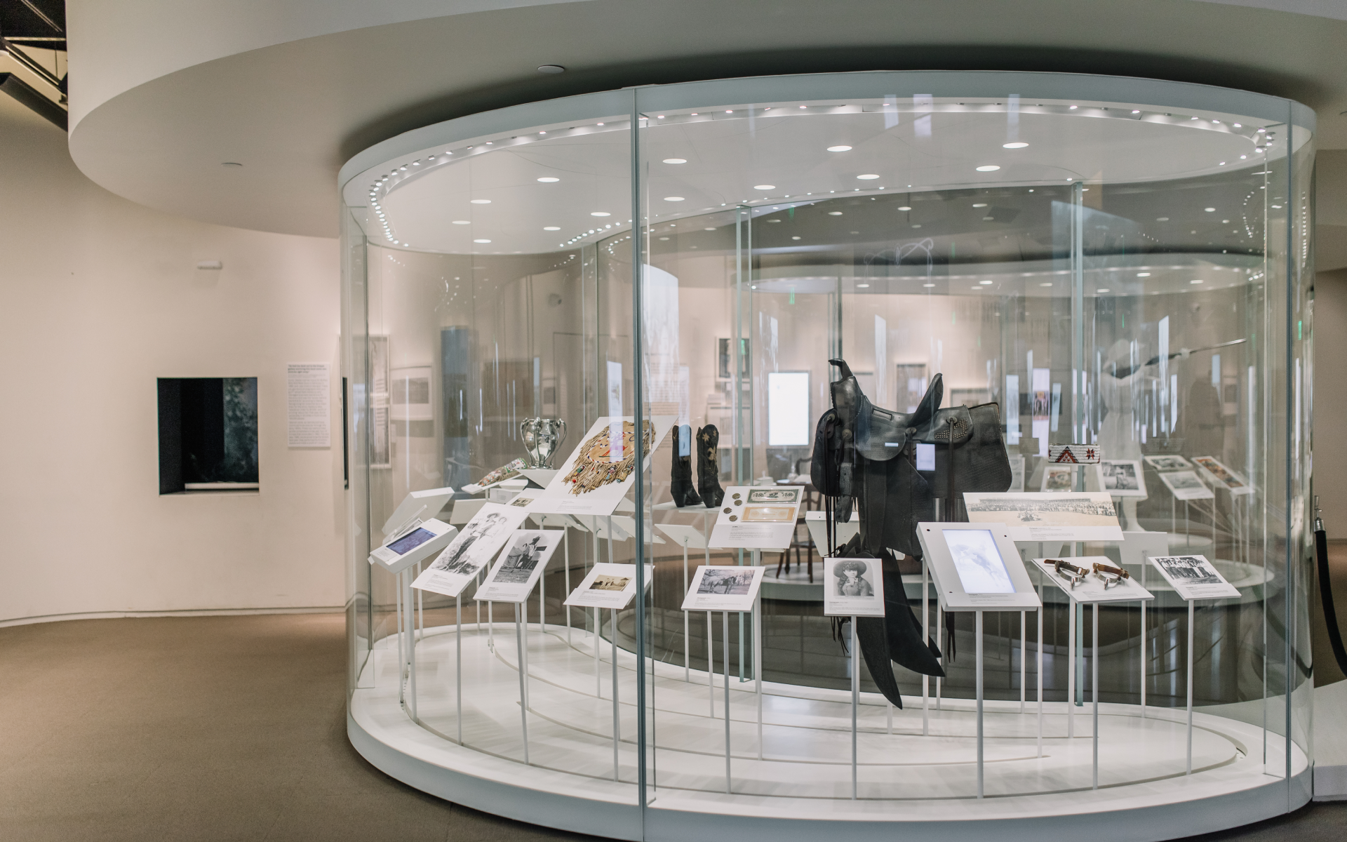 A modern museum exhibit with artifacts, photos, and digital displays in a circular glass case, set in a bright, white gallery space.