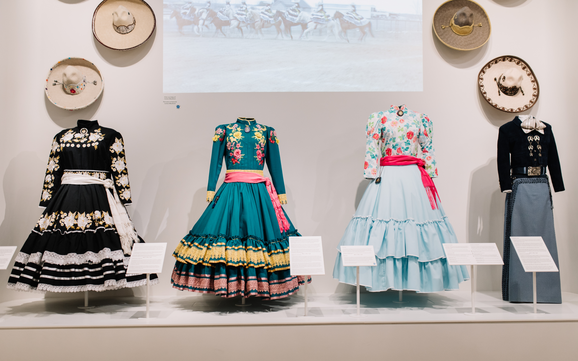 Four traditional Mexican dresses are displayed on mannequins against a white wall adorned with sombreros. The colorful dresses feature floral embroidery, ruffles, and sashes, reflecting cultural heritage.
