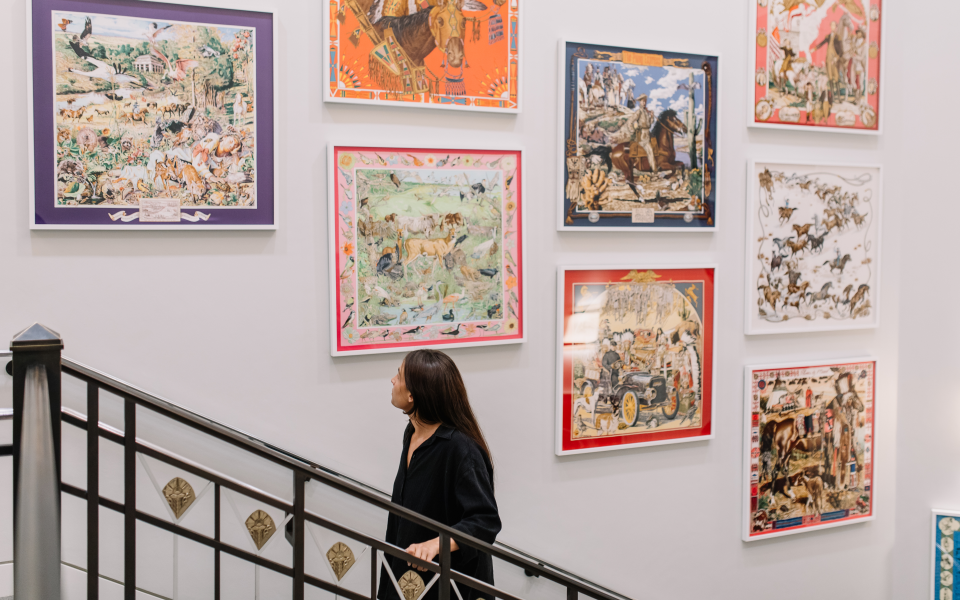 A woman with long dark hair walks up a staircase, looking at colorful framed art pieces depicting various scenes on a white gallery wall.