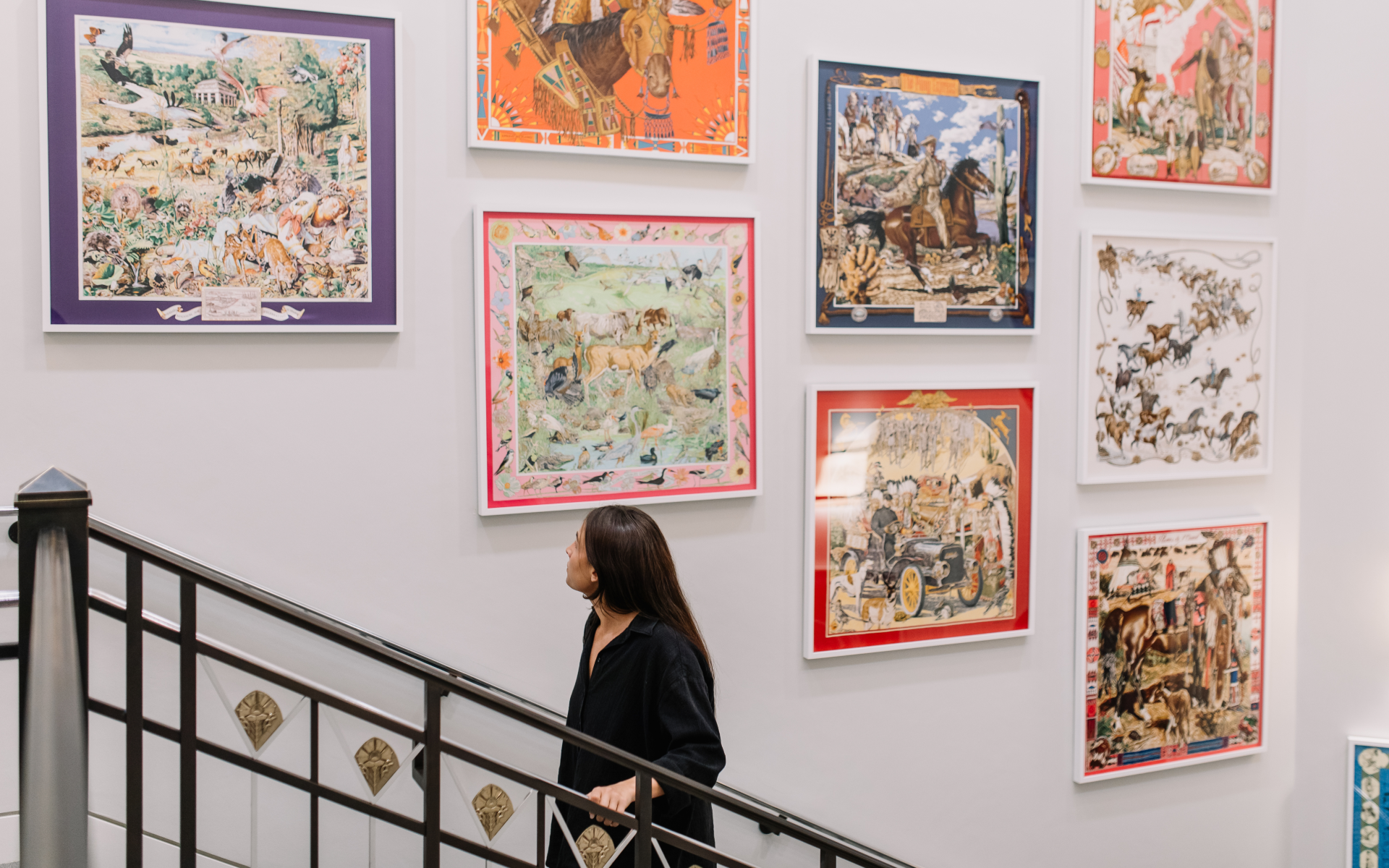 A woman with long dark hair walks up a staircase, looking at colorful framed art pieces depicting various scenes on a white gallery wall.