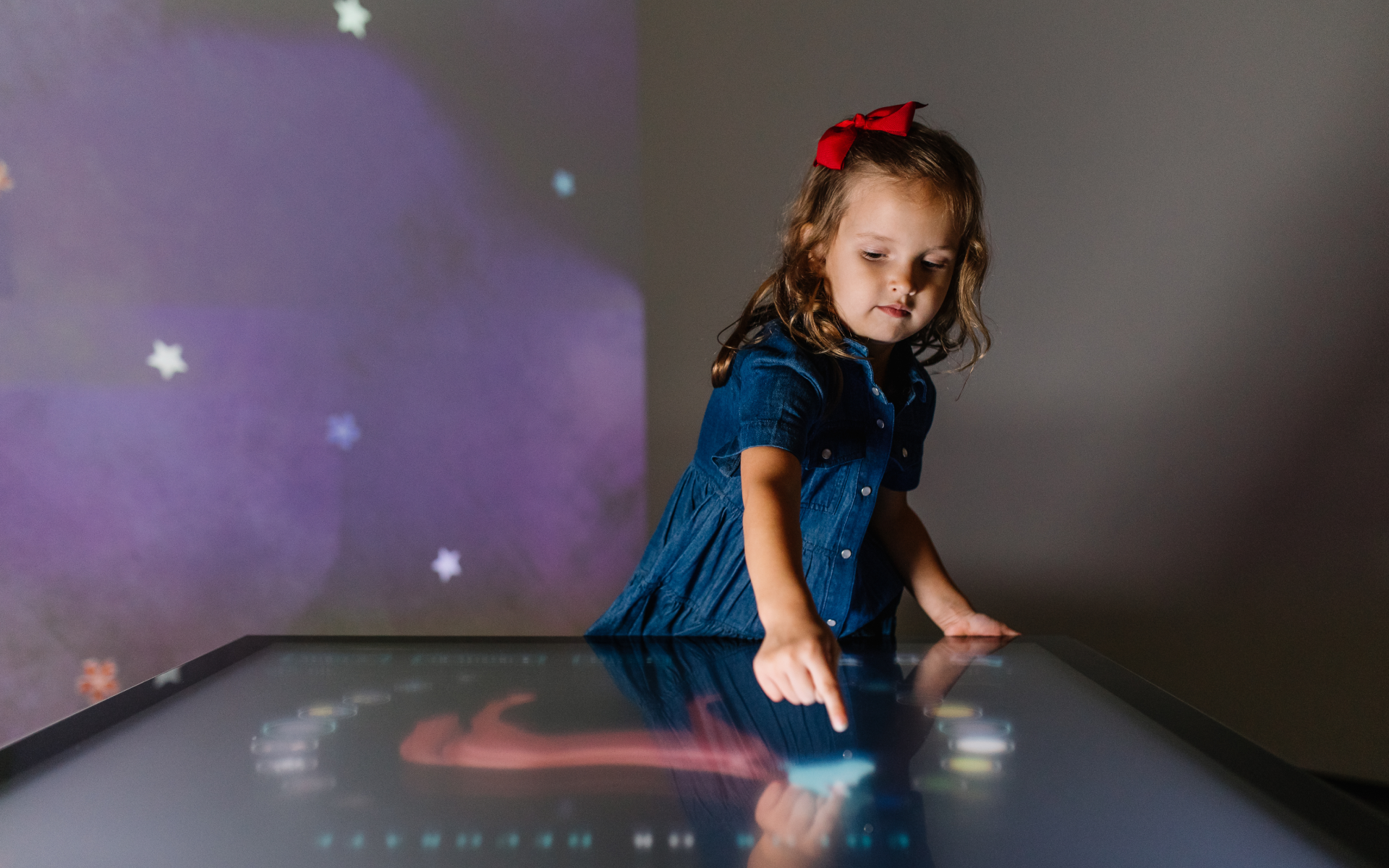 A young girl in a blue dress with a red bow uses a large interactive touchscreen table, with stars projected on the purple wall behind her.
